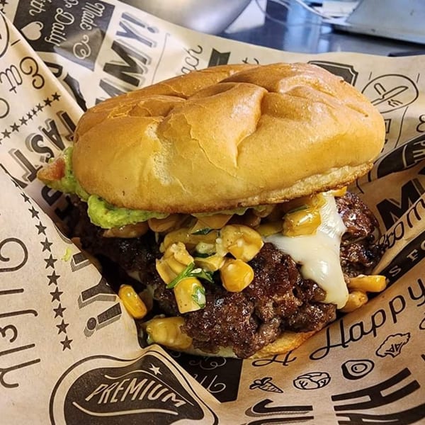 A Poppy's Smokehouse burger with melted cheese, corn, and guacamole, served on branded paper.
